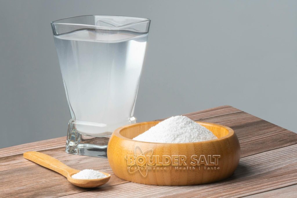 Wooden bowl of Boulder Salt with a small spoon and a cloudy glass of water on a wood table.